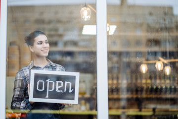 woman stands and opens a wide sign through the shop window and smile.