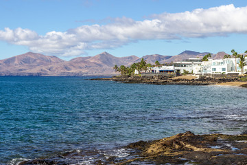 Fototapeta na wymiar Rocky coast of Puerto del Carmen at Canary island Lanzarote with lava rocks and blue water in the foreground, white houses, palm trees and volcanic mountain range in the background