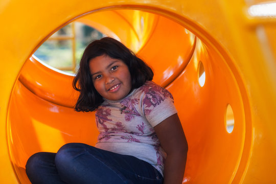 A cute latino girl with short hair playing in a kids playground, sitting inside a circular tube slide.