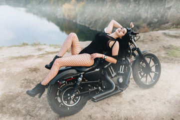 Beautiful brunette girl with long hair in a black dress and stockings seductively lies on an expensive motorcycle against the backdrop of nature in foggy weather. Portrait of a sexy woman on a bike.
