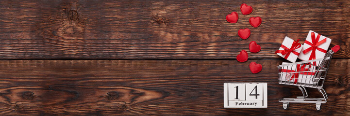 Shopping trolley, gifts, hearts on brown wooden background. 14 February. Valentine's Day. Extra wide panorama banner background