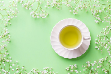 Green tea in a white cup with gypsophila flowers on green pastel background. Top view. Copy space.