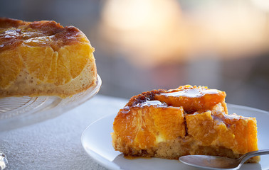 Piece of mandarin and honey sponge cake on white plate with a partial view of the sponge cake on an unfocused landscape background.