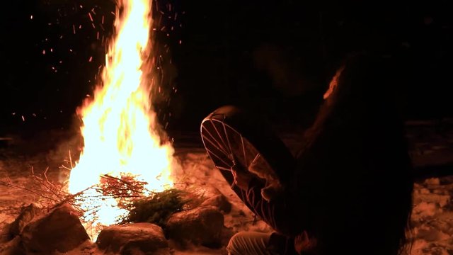 A spiritual native man is seen from behind by night, sitting by a burning fire and playing a sacred drum to the spirit world during magical ceremony