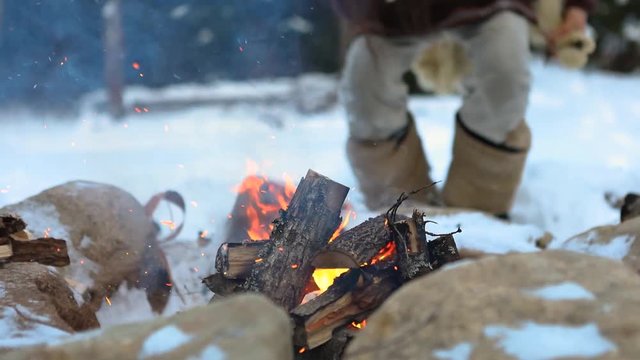 Close up and selective focus footage of a burning camp fire with snow on ground. as a blurry native man wearing cherokee style clothes sits for warmth