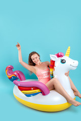 Summer Freestyle. Young woman with freckles isolated on blue sitting on swim ring unicorn shape hand up smiling cheerful