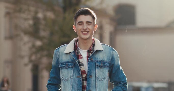 Portrait shot of the young Caucasian handsome guy student in the blue jeans jacket standing at the street and smiling to the camera. Outdoor.