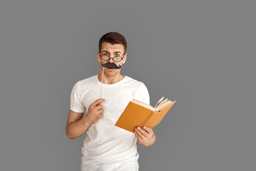 Freestyle. Young student in eyeglasses standing isolated on grey with book and party props mustache smiling confused