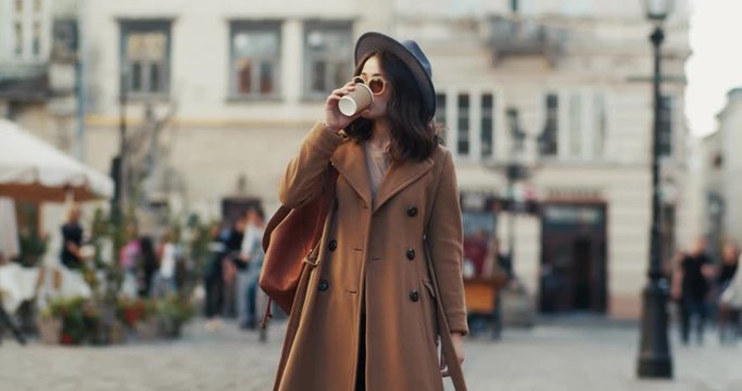 Stylish Caucasian young beautiful woman, model or a female single traveller in a hat and cout having a walk at the town and looking at the sides while smiling and drinking coffee to-go. Outdoors.
