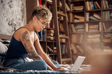 Side Hustle. Young woman short hair in glasses sitting on bed working online on laptop joyful