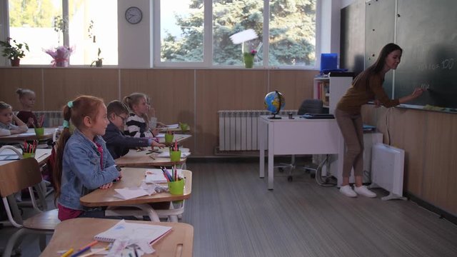 Clever preadolescent redhead girl raising hand and going to blackboard during math lesson in primary school. Mixed race female teacher writing math task on blackboard for elementary school students