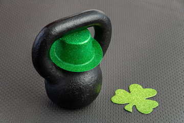 Naklejka na ściany i meble Black iron kettlebell with green leprechaun hat on a black gym floor, holiday fitness, green shamrock