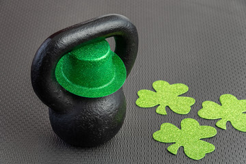 Naklejka na ściany i meble Black iron kettlebell with green leprechaun hat on a black gym floor, holiday fitness, green shamrocks