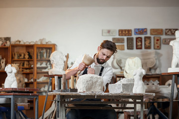 Bearded craftsman works in white stone carving with a chisel. Creative workshop with works of art.
