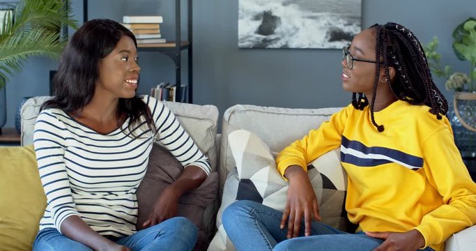 Two African American young girls, best friends talking and smiling while sitting on the couch, gossiping and sharing secrets. At home.