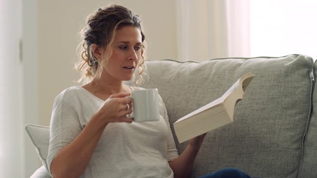 Woman Reads Book And Sips From Mug While Reclining On Couch With Feet Up