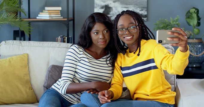 Portrait shot of the two young African American friendly girls smiling to the camera while taking selfie photos on the smartphone and sitting on the sofa at home.
