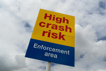 Bright yellow and blue high crash risk enforcement area warning sign against a cloudy blue sky.