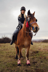Young caucasian blonde woman female girl on the horse riding in nature wearing helmet in winter or autumn day against a a gray sky in the field