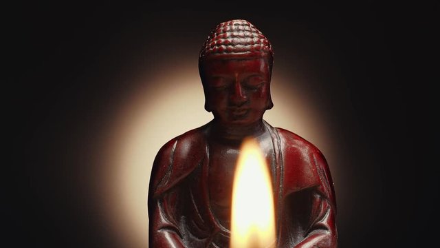 Panning shot alongside sitting in meditation statue of Buddha. Burning candle on foreground. Dolly camera move, slowly sliding, close-up shot