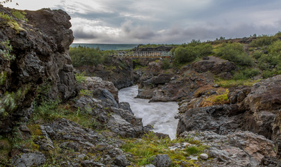 View of a mountain river that flows in a small gorge among volcanic rocks. The bridge over the river in the background
