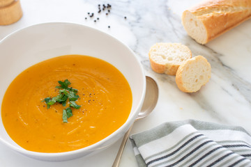 Pumpkin soup in white bowl with bread and garnished with chopped parsley and cracked pepper on white marble background