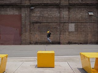 Asian boy wearing a yellow hat jumping infront of an old warehouse brick wall. Foreground also have three pieces of yellow street furniture.