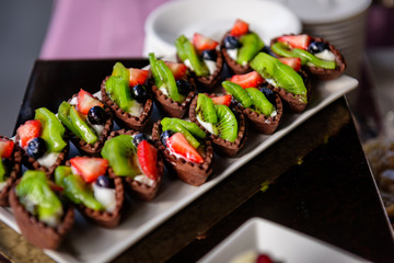 Delicious sweet tartlets made with chocolate dough with fresh fruit and berries close-up. Small cakes with strawberries, kiwi, grapes. Banquet service at wedding, anniversary.