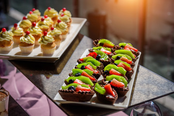 Cakes with fresh fruit and berries on the holiday table, close-up. Cupcakes with cream. Sweet table at gala dinner.
