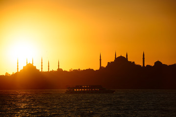 Naklejka premium Sun setting over Blue Mosque with Hagia Sophia in silhouette on the Bosphorus with boat Istanbul