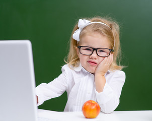 Cute little girl in glasses is working on a laptop at school and showing displeased funny faces