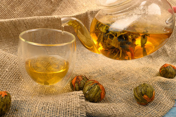 A ball of flower tea brewed in a glass teapot, a glass of tea and several balls of flower tea on a background of rough homespun fabric. Close up.