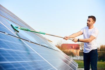 Young man is washing a solar panel with a mop, pv plant in rural area, cleaning increases to high...