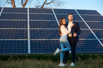 Young and beautiful couple is standing close to each other by a solar panel, man shows thumb up,...