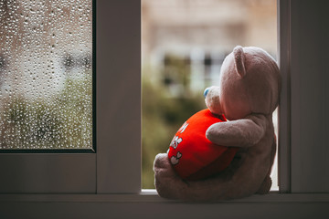 A toy pink sad missing bear with a red plush heart sitting on the windowsill. Autumn rainy day....