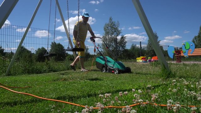 Caucasian man cut grass with lawn mower near swing in playground. Gimbal motion
