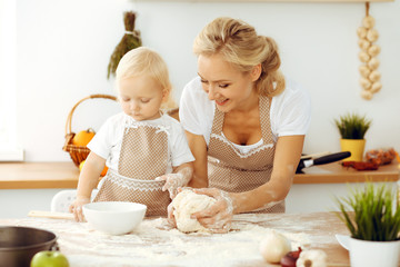 Little girl and her blonde mom in beige aprons playing and laughing while kneading the dough in kitchen. Homemade pastry for bread, pizza or bake cookies. Family fun and cooking concept