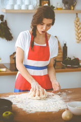 Young brunette woman cooking pizza or handmade pasta in the kitchen. Housewife preparing dough on wooden table. Dieting, food and health concept