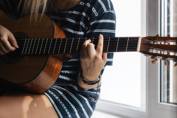 The young woman in the striped t-shirt sitting on the windowsill and playing acoustic guitar. Close...