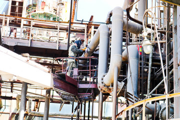 Worker repairman fitter welder repairs a pipeline at a refinery petrochemical chemical industrial plant. A welder in a cradle is cooking a pipe