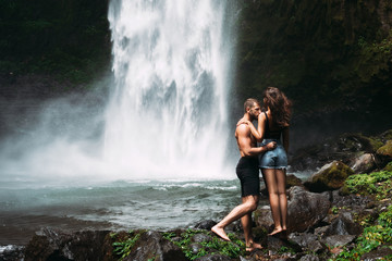 Fototapeta na wymiar A man of athletic build kisses a beautiful girl at the waterfall. Honeymoon trip. Declaration of love. A couple in love on a waterfall. Happy couple in Bali. Beautiful couple travels the world