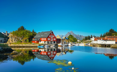 Village on the shore of the fjord, Lofoten Islands, Norway. Sunny day, clear blue sky, colorful house, reflection in the water, forest and mountains in the background. - obrazy, fototapety, plakaty