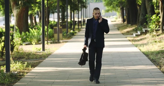 Successful young man has good news on mobile phone and shows joyful pose walking along shadow city street in summer