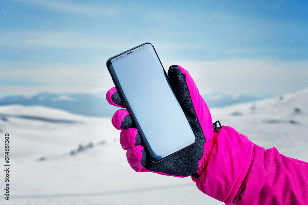 Poster phone in hand with glove on snowy mountain peak. the concept of using a mobile phone to follow a hik