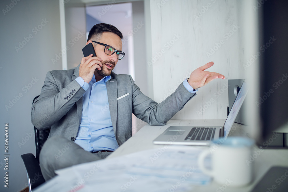 Wall mural handsome classy unshaven caucasian businessman in suit and with eyeglasses sitting in his office and