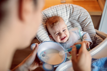 Hungry crying baby with bib sitting in his chair and waiting for his mother to feed him with delicious porridge.