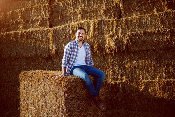 Handsome caucasian bearded smiling farmer in plaid shirt and jeans sitting on bale of hay while looking at camera. © Dusan Petkovic
