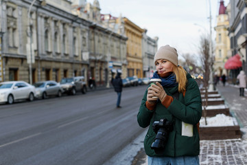 Girl in winter clothes with camera and map in the historical center of the city is holding a cup of coffee on the street