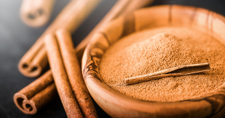Cinnamon powder in wooden bowl on dark stone table. Cinnamons sticks on black board.