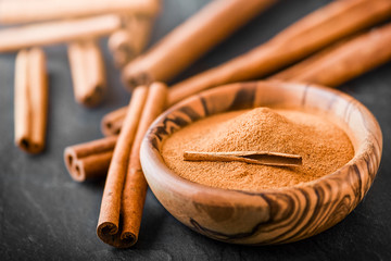 Cinnamon powder in wooden bowl on dark stone table. Cinnamons sticks on black board.